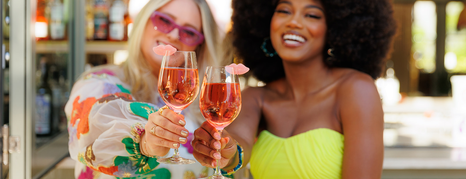 two women in bright and colorful dresses smiling and holding a cocktail