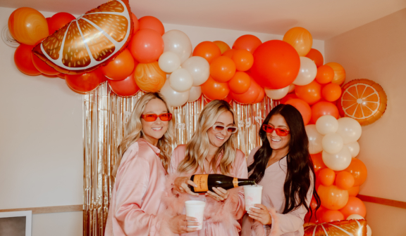 three women with vibrant orange balloons in background pouring champagne